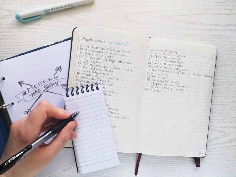 A hand holding a pen ready to take notes with a reading log of Agathie Christie books in the background