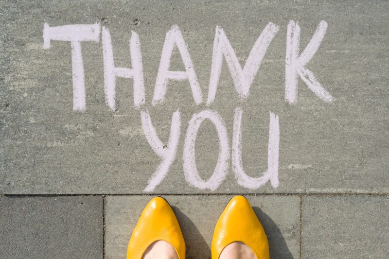 A woman's feet in bright yellow shoes on a sidewalk with a message that says Thank You written in chalk