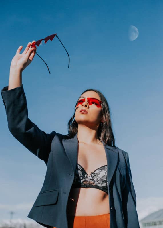 A model wearing a blazer and a bra holds up sunglasses in the air, with a blue sky and moon in the background