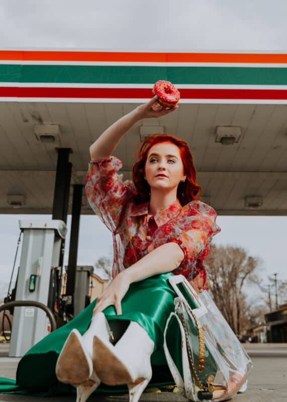 a model with red hair and a colorful outfit sits outside a 7-11 holding a doughnut in the air. Photography and styling on a budget can be a fun creative outlet for adults