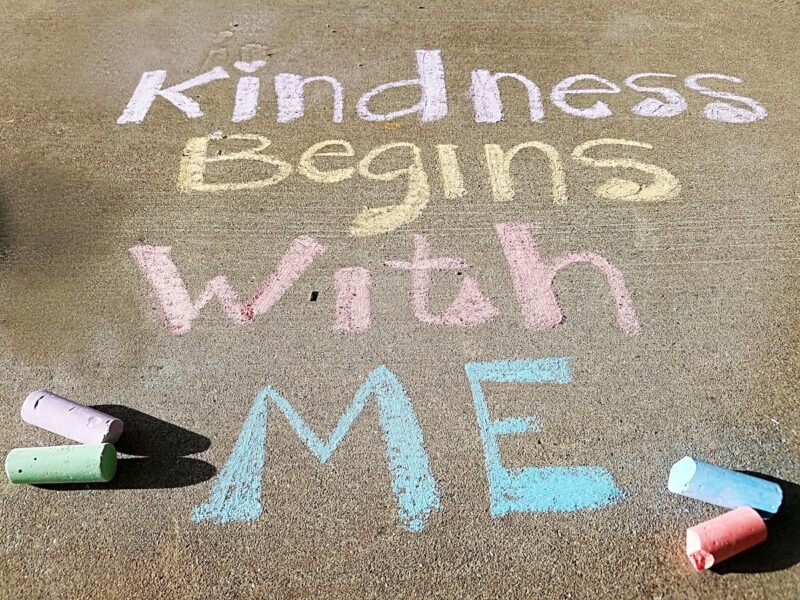 Colorful message written in chalk on a sidewalk that says "Kindness Begins with Me." Positive thinking exercises in your journal can help you practice self kindness