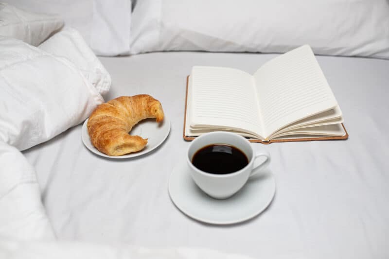 Croissant, journal and coffee in a bed with white linens. When practicing morning journaling, morning journal prompts can help get started