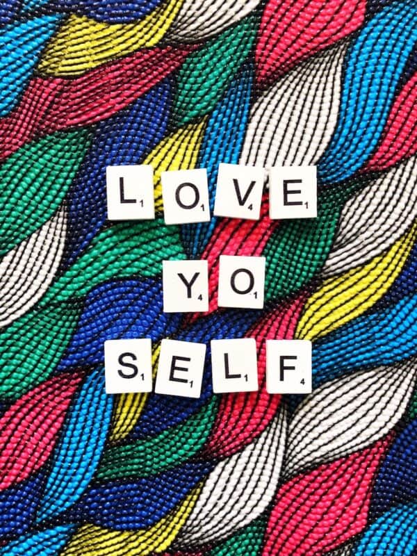 Scrabble tiles spell out "Love Yo Self" against a colorful multicolored textured beaded background